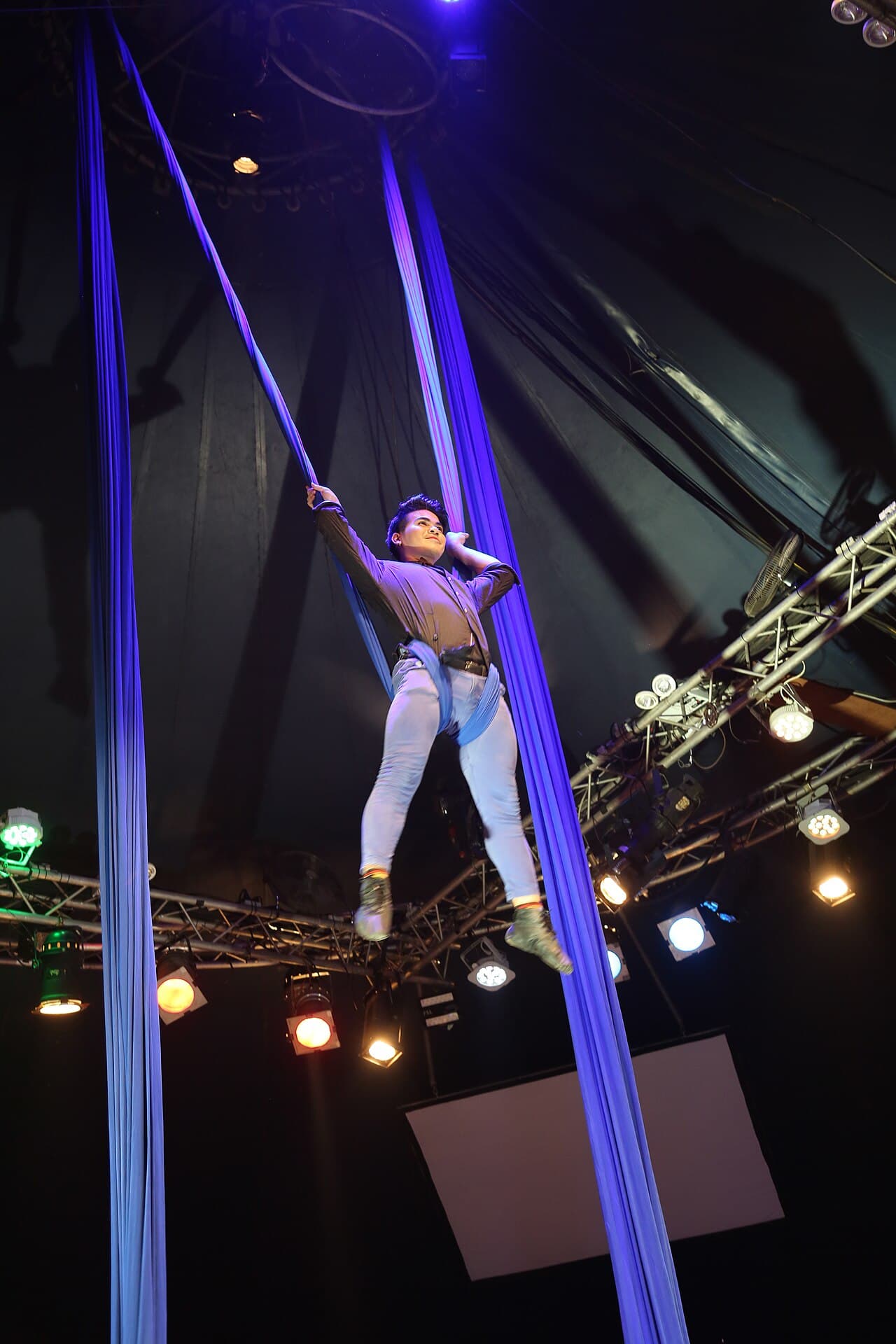 A Phare aerialist part way up the silks, with the big top's rigging and stage lights behind.